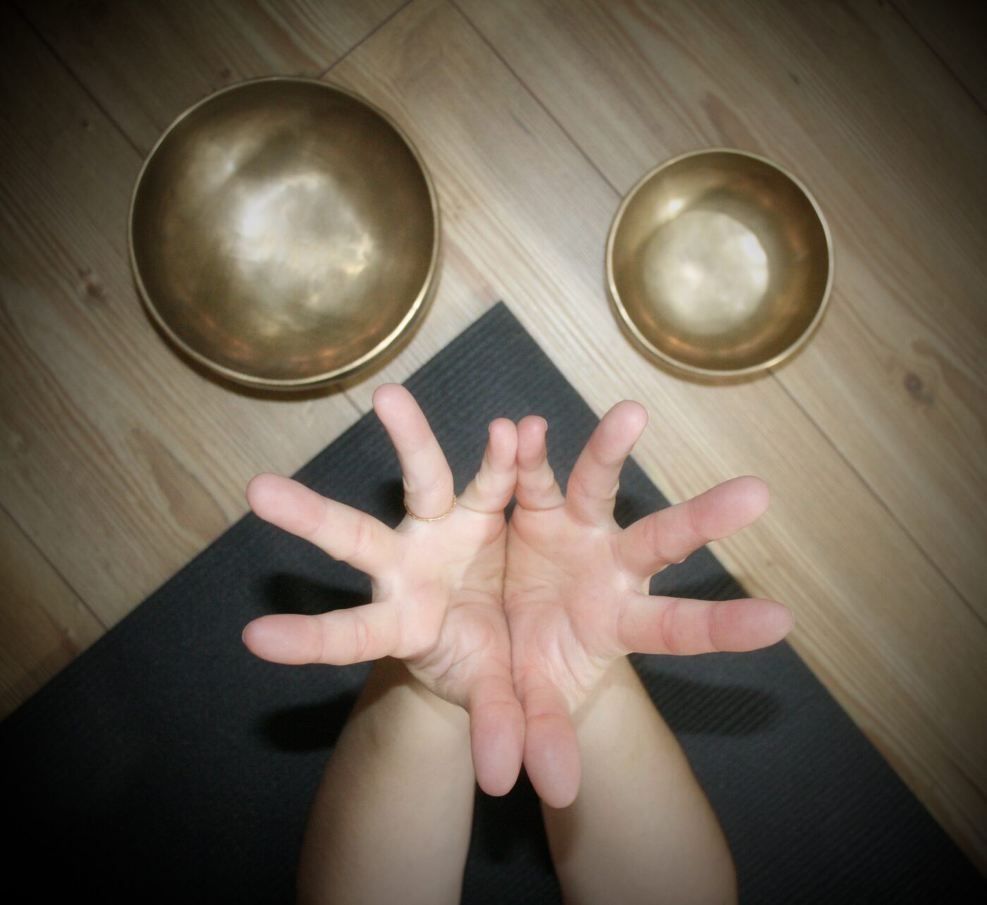 Hands are reaching directly up to the camera, with a yoga mat and two Tibetan singing bowls shown on the floor below