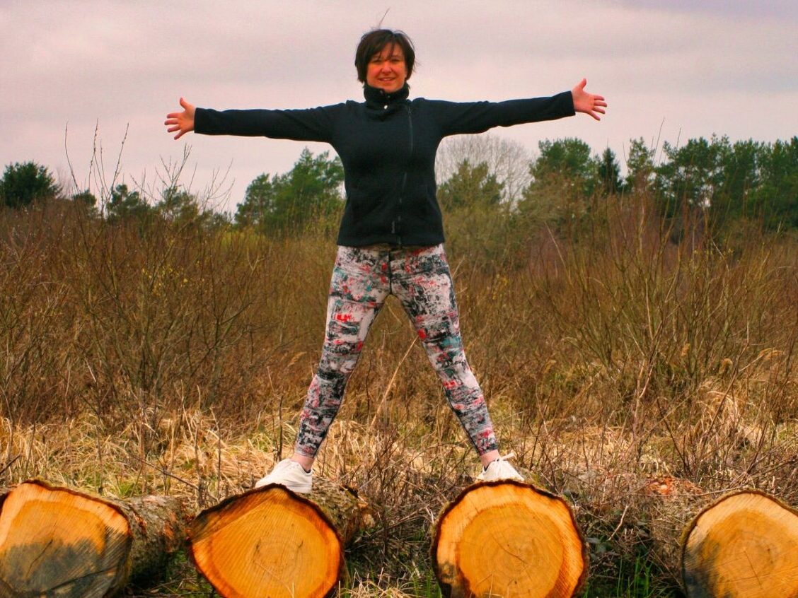 Liz is outdoors and is balancing on large cut logs in a star posture with open arms